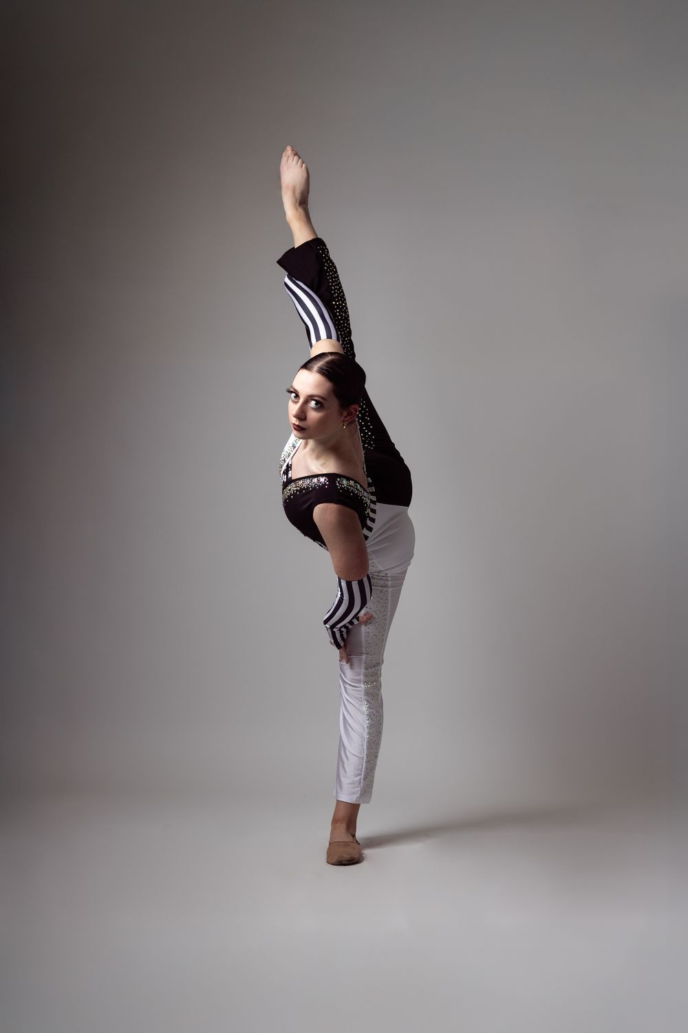 A flexible dancer balances on one leg while raising the other high, wearing white pants and a black top in a studio.