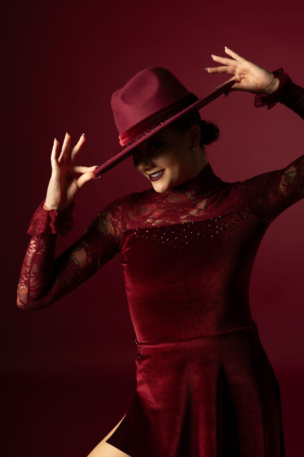 A woman in a burgundy lace dress and matching wide-brim hat poses dramatically against a deep red background.