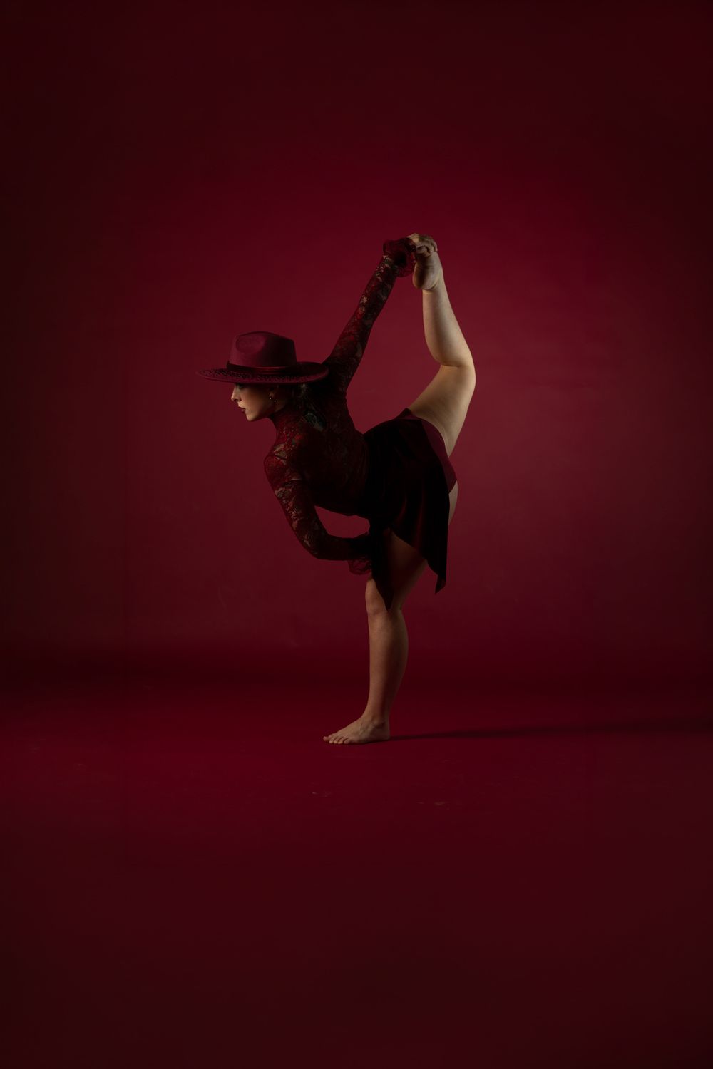A flexible dancer in a red outfit and hat performs a standing split against a deep red background.