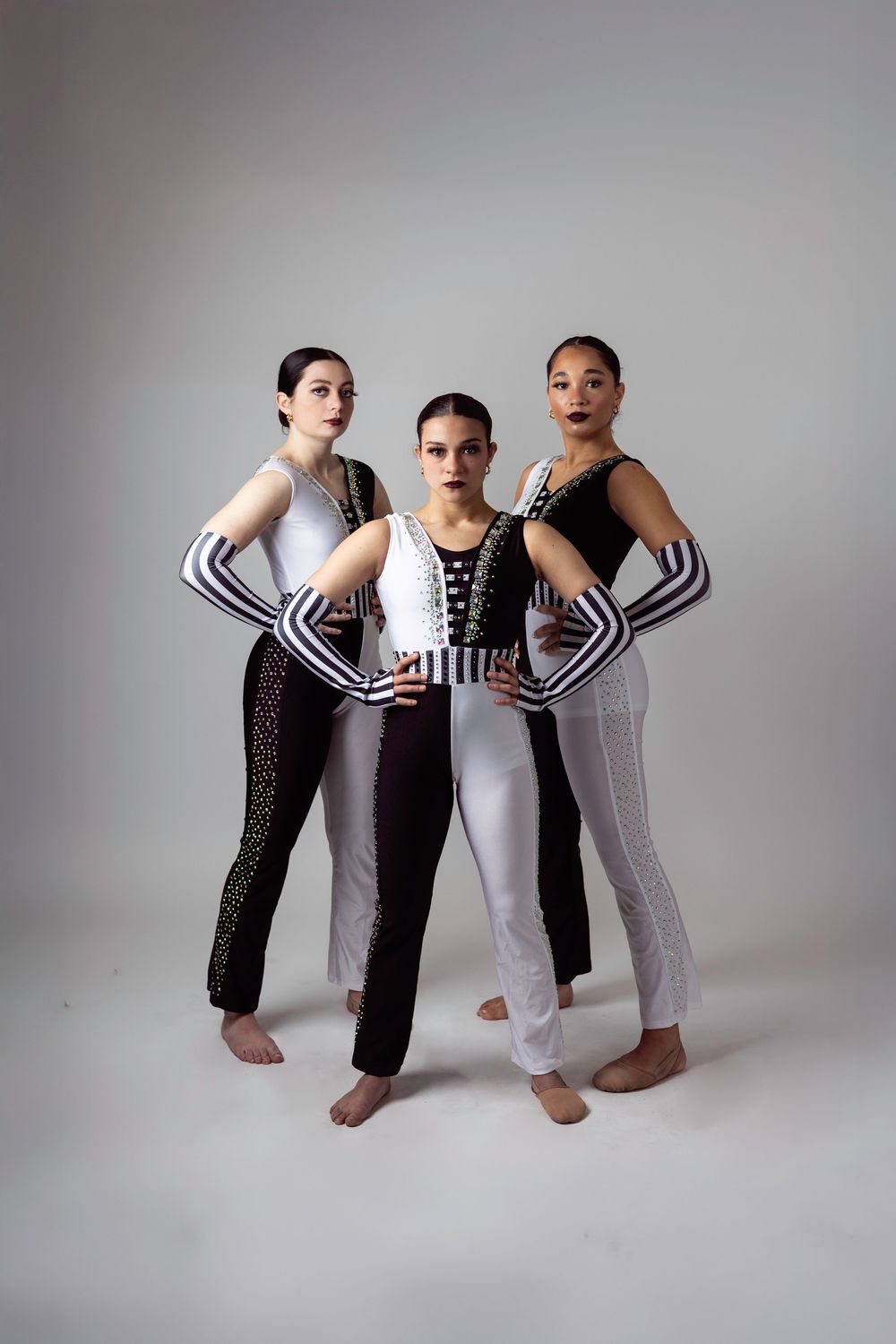 Three dancers in black and white costumes pose confidently barefoot against a light gray studio background.