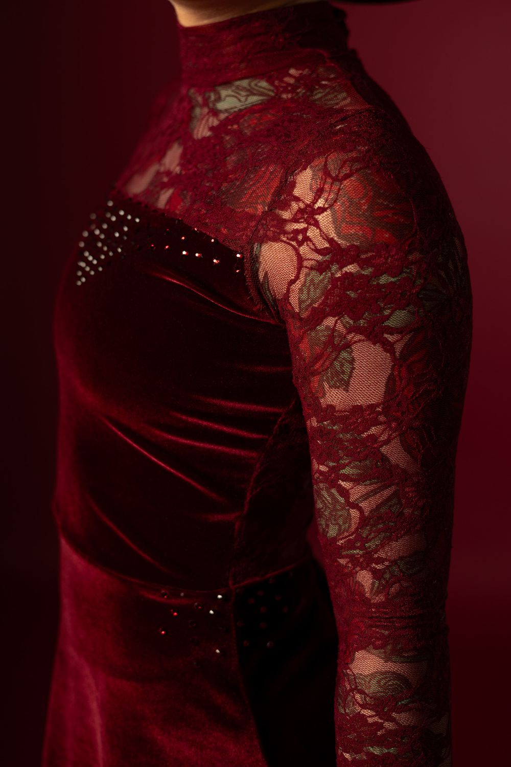 Close-up of a burgundy velvet and lace long-sleeve dress with rhinestone details on a dark red background.