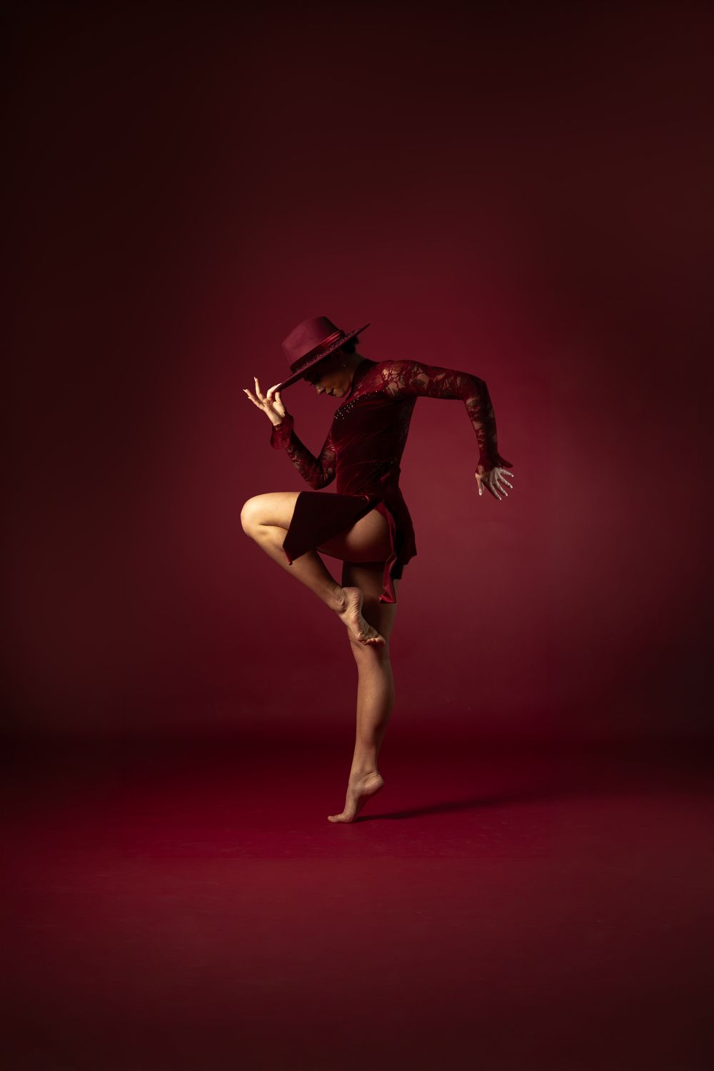 A dancer in a red outfit and hat performs a dynamic pose on one leg against a dark crimson background.