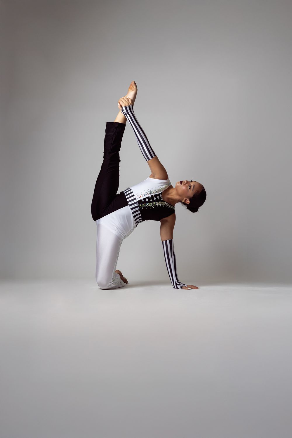 A flexible gymnast in black and white outfit performs a graceful backbend split pose on a gray studio background.