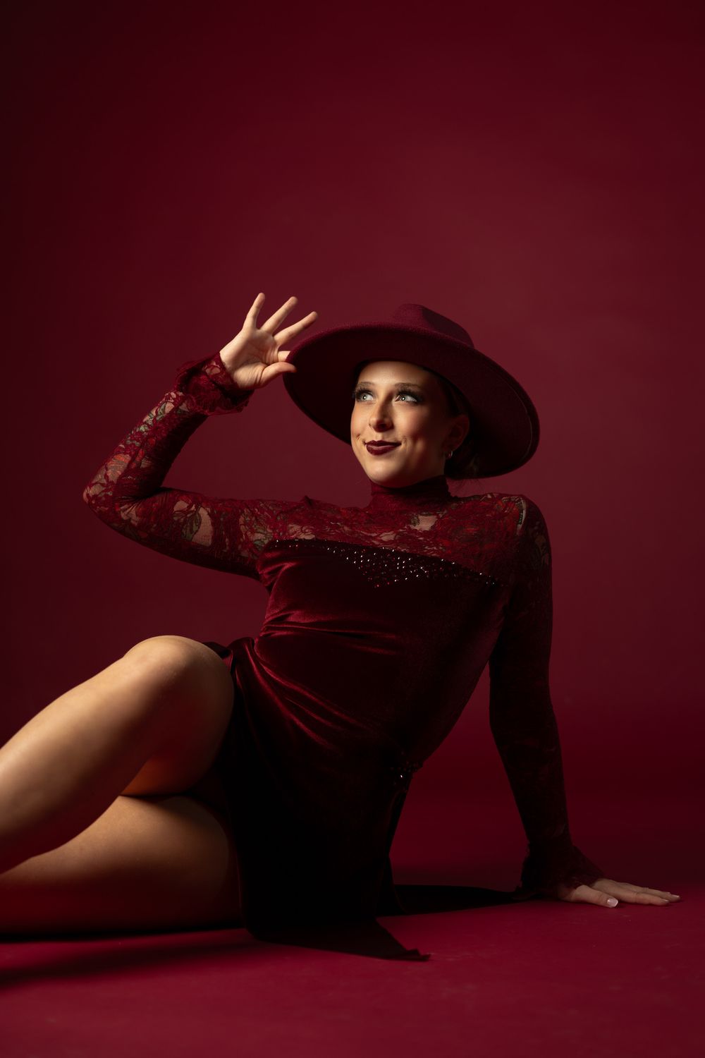 Woman in burgundy velvet top and wide-brim hat poses elegantly on a dark red studio floor.