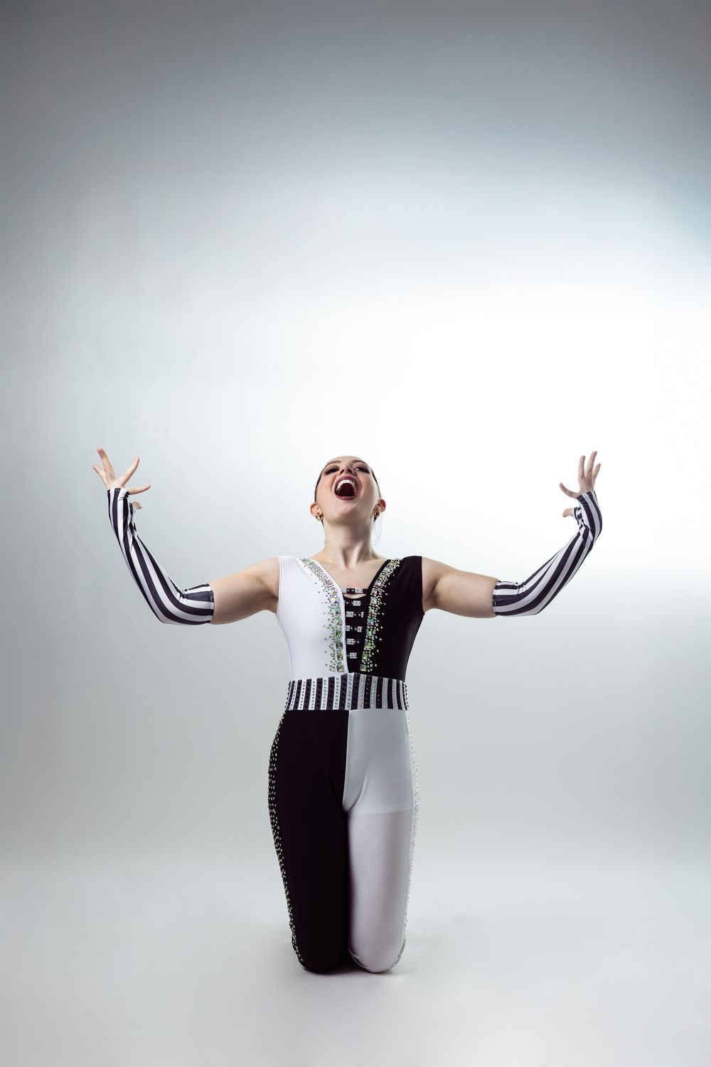 A dancer in a black and white costume kneels dramatically with arms raised and mouth open on a gray background.