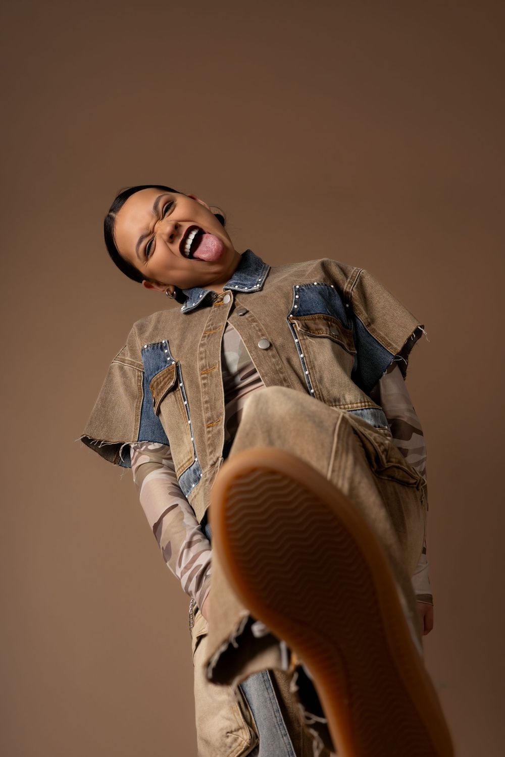 A laughing person in a denim jacket kicks their foot toward the camera against a brown background.
