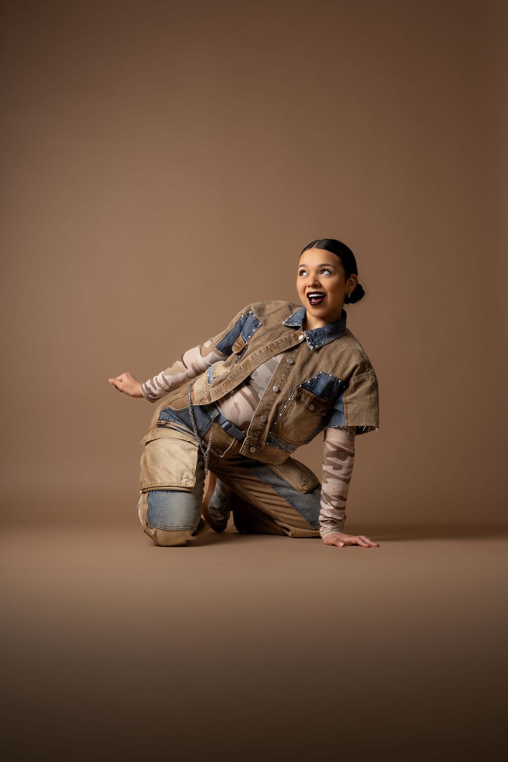 Woman in distressed denim outfit poses on floor against brown background, smiling confidently.