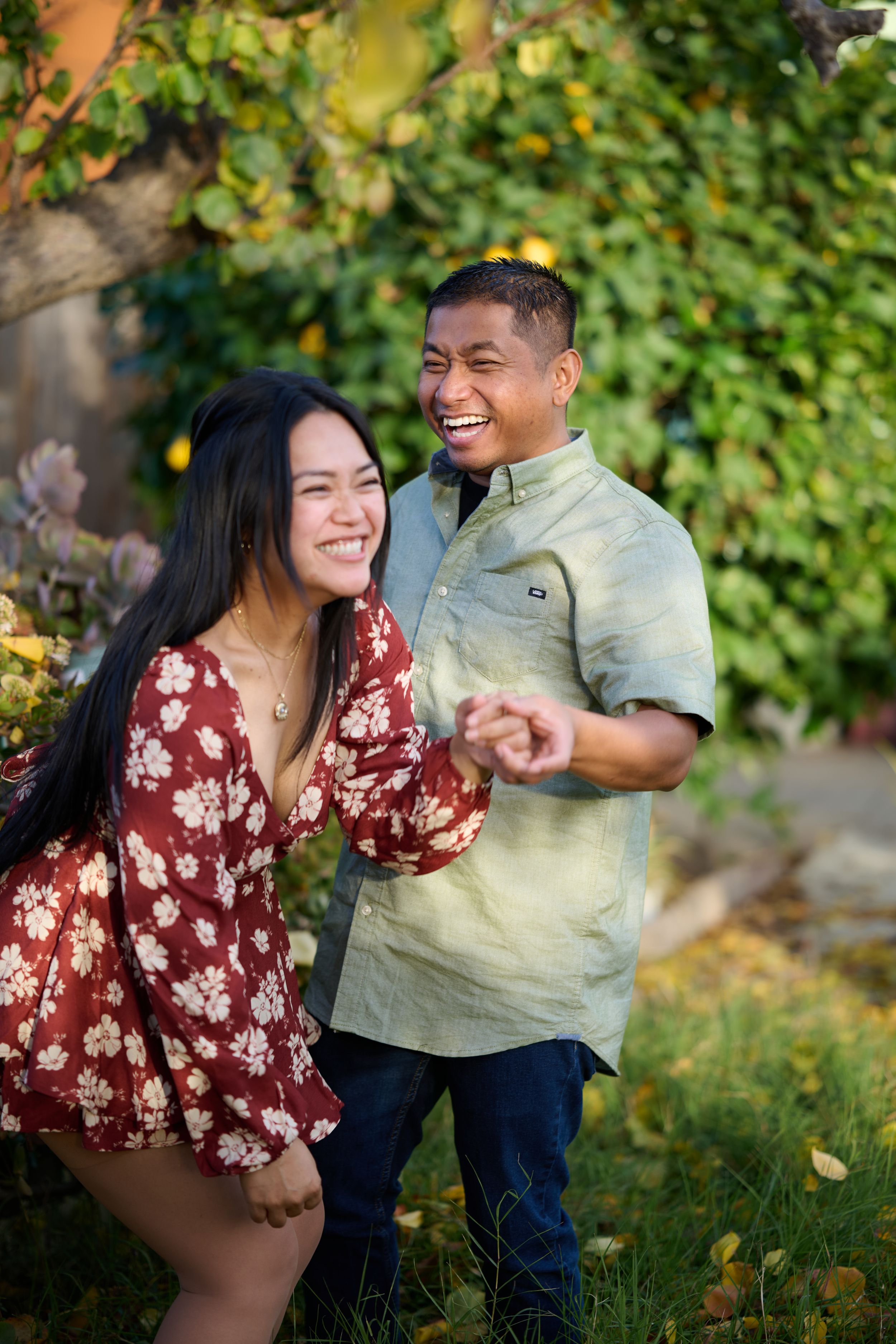 A couple laughs together outdoors in front of green foliage, wearing floral red dress and light green shirt.