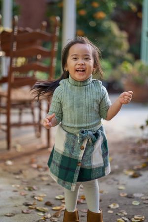 Young child wearing green sweater and plaid skirt jumps joyfully among fallen autumn leaves outdoors.