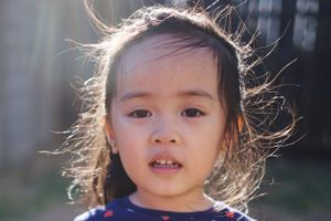 A child with windswept hair backlit by sunlight, wearing a blue shirt while looking at the camera.