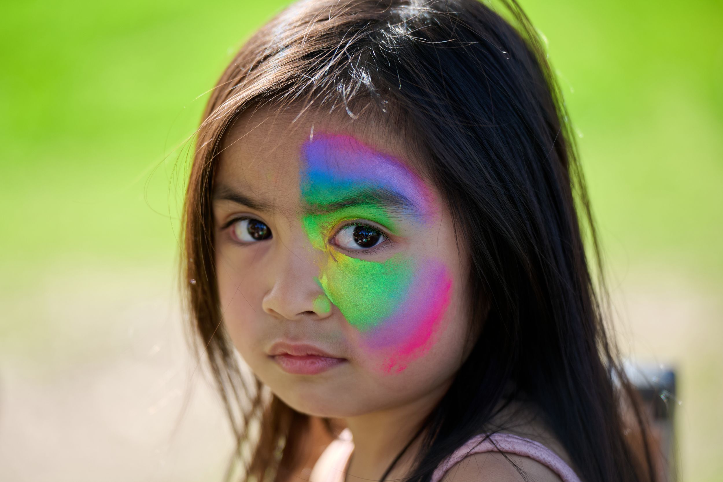 A colorful rainbow pattern painted on a side of a person's face against a bright green outdoor background.