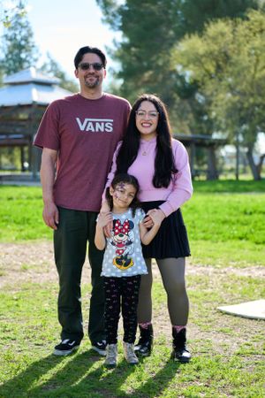 A family poses together on a sunny day in a grassy park area, with trees in the background.