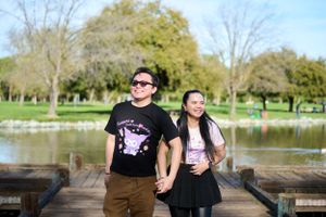Two people hold hands while standing on a wooden pier by a tranquil pond in a lush park setting.