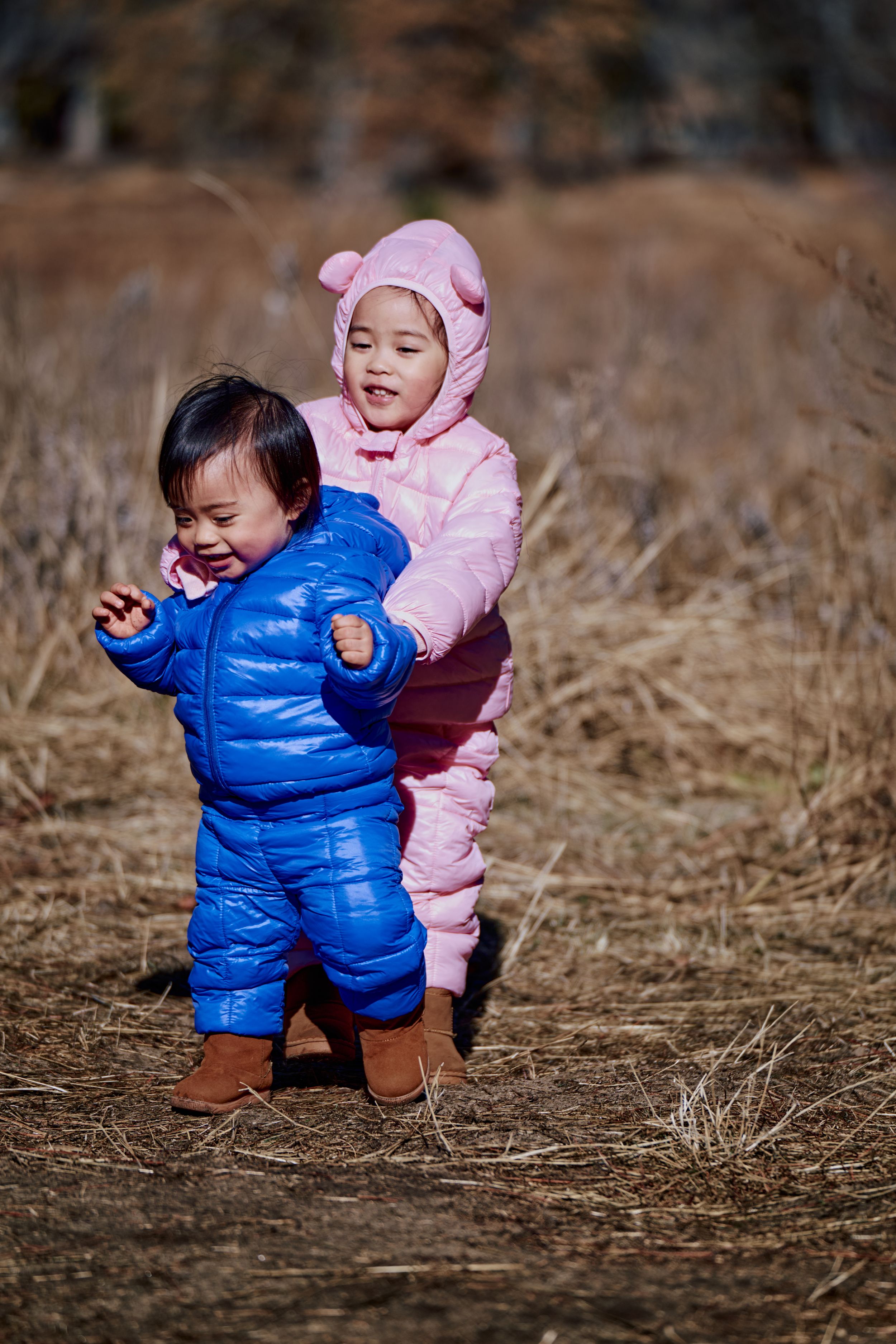 Two small children in puffy winter coats, one pink and one blue, playing together in a field of dry grass.
