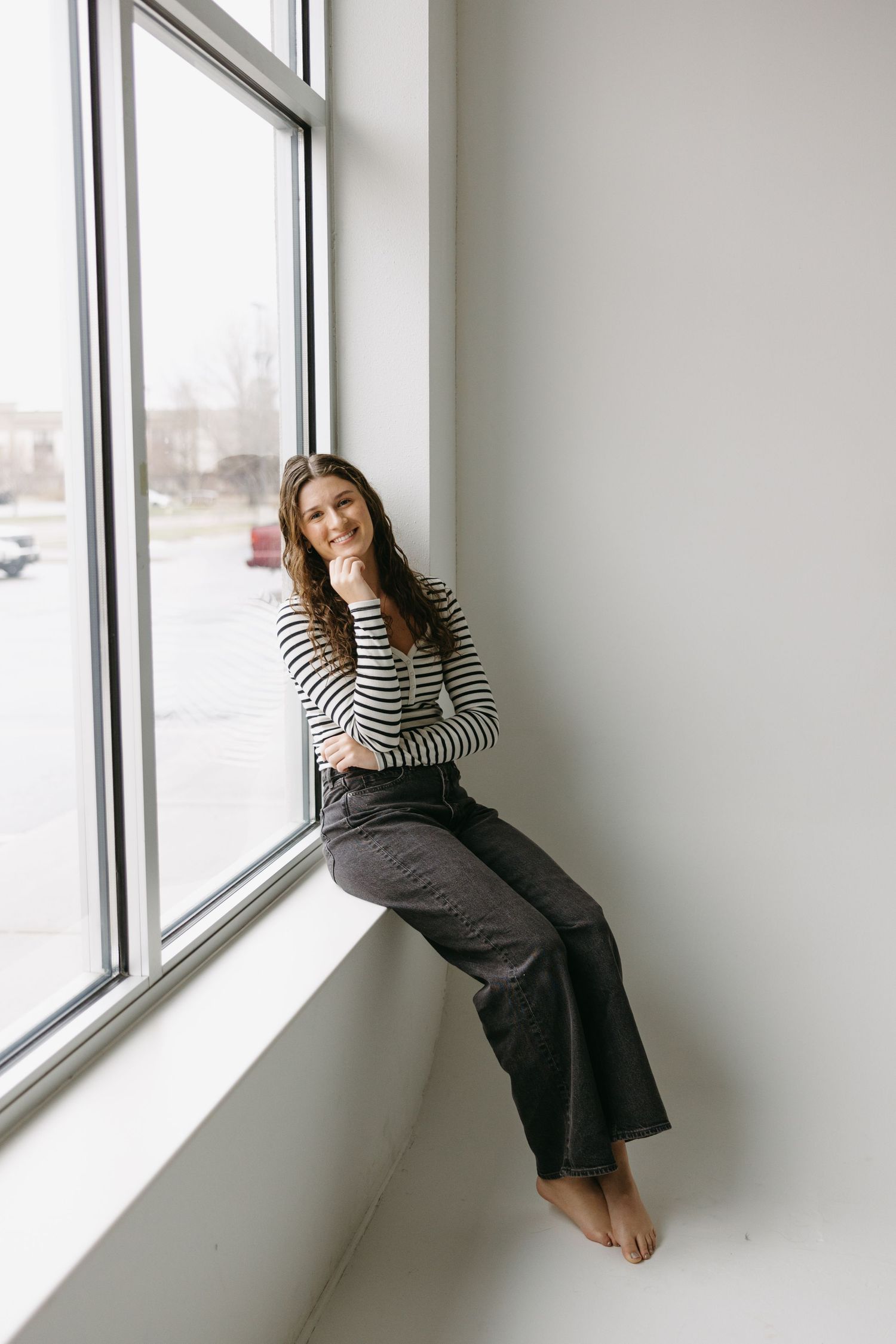 Relaxed indoor portrait by window wearing striped top and dark pants in natural light setting.