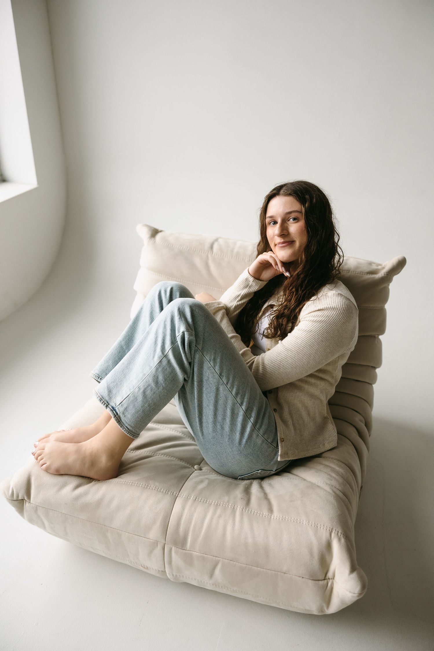 Lifestyle portrait on beige cushion wearing cream sweater and light denim in bright white room.