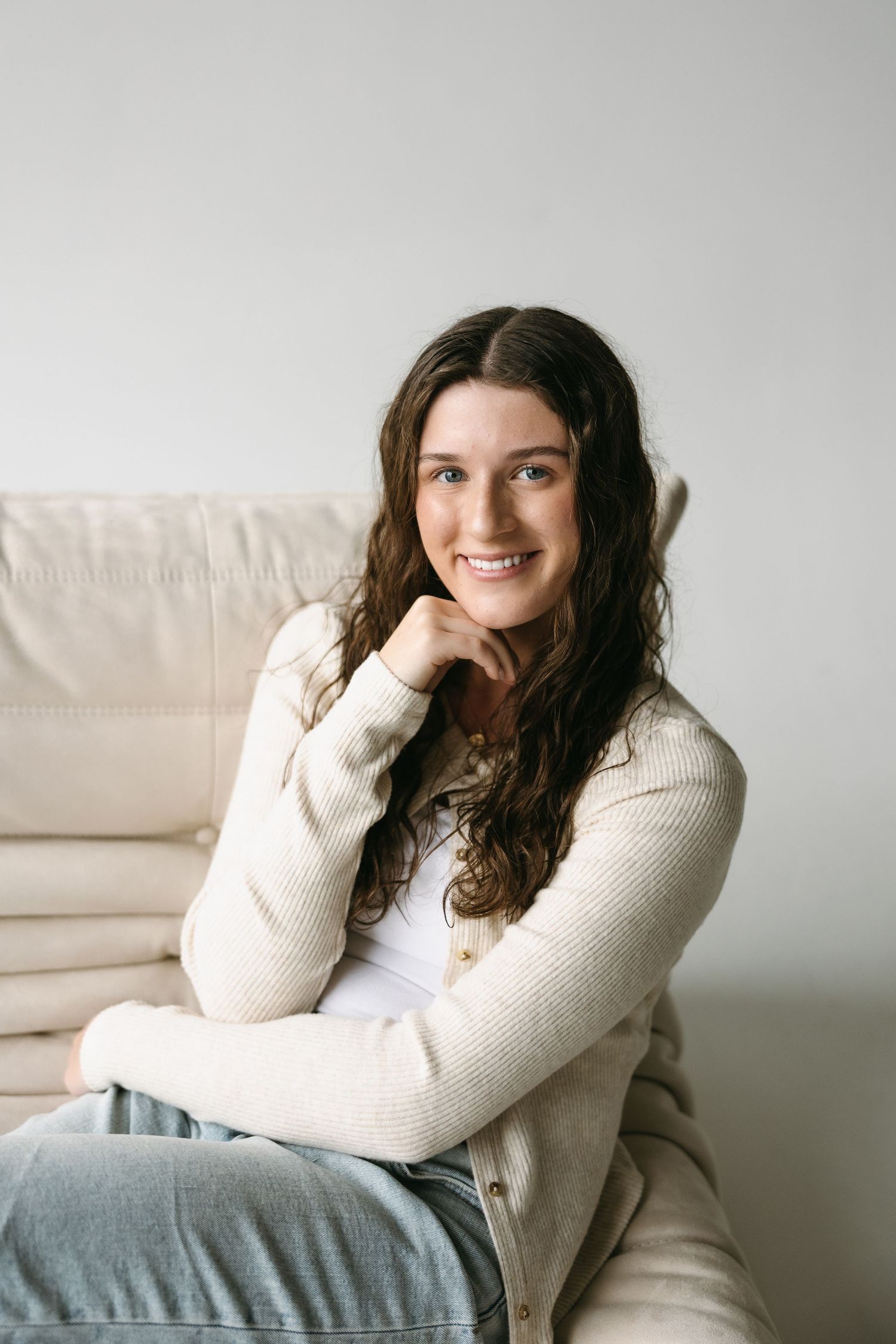 Natural lifestyle portrait on beige couch wearing cream cardigan and light denim in bright setting.