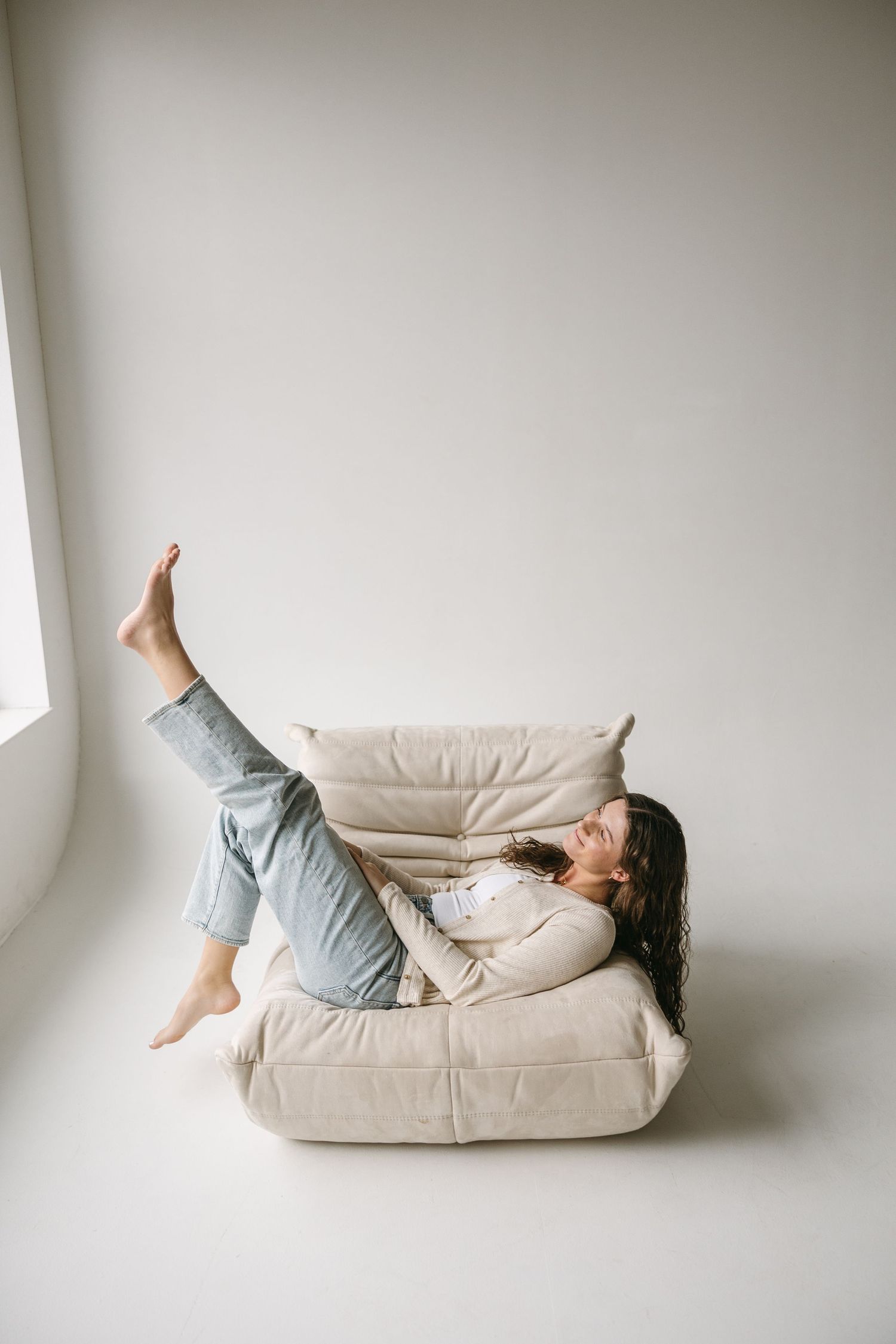 Playful lifestyle portrait lying on beige floor cushion with leg raised against white wall.