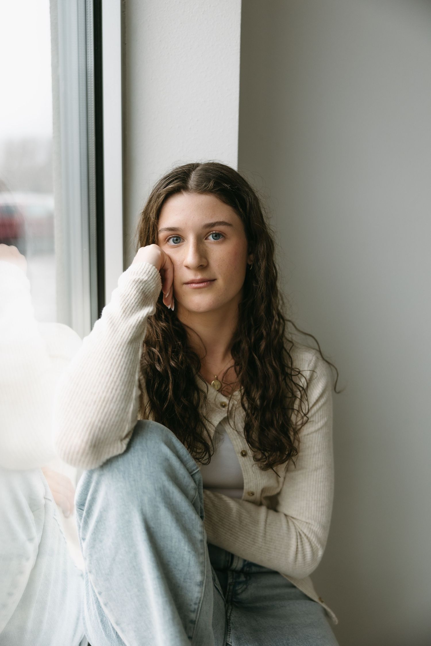 Natural light portrait by window wearing cream sweater and light denim in minimalist setting.