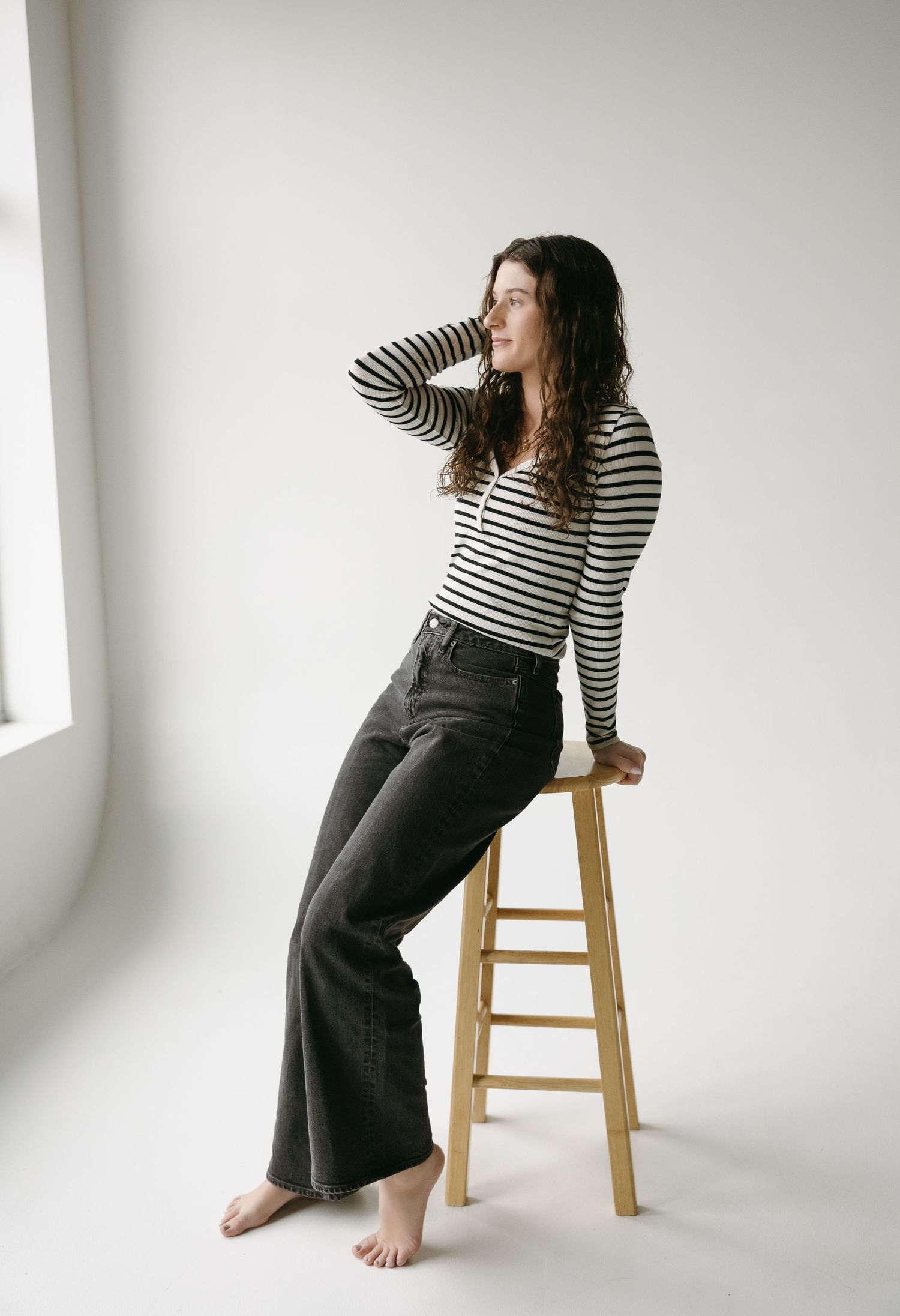 Casual lifestyle photo of person in striped shirt sitting on wooden stool by window.