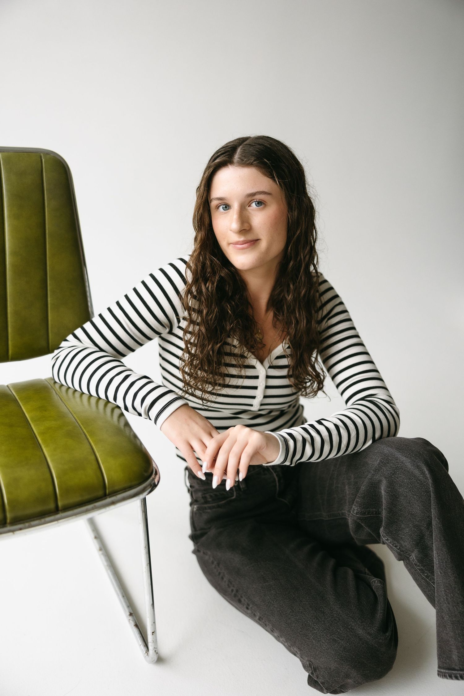 Casual portrait in black and white striped sweater sitting beside olive green velvet chair against white background.