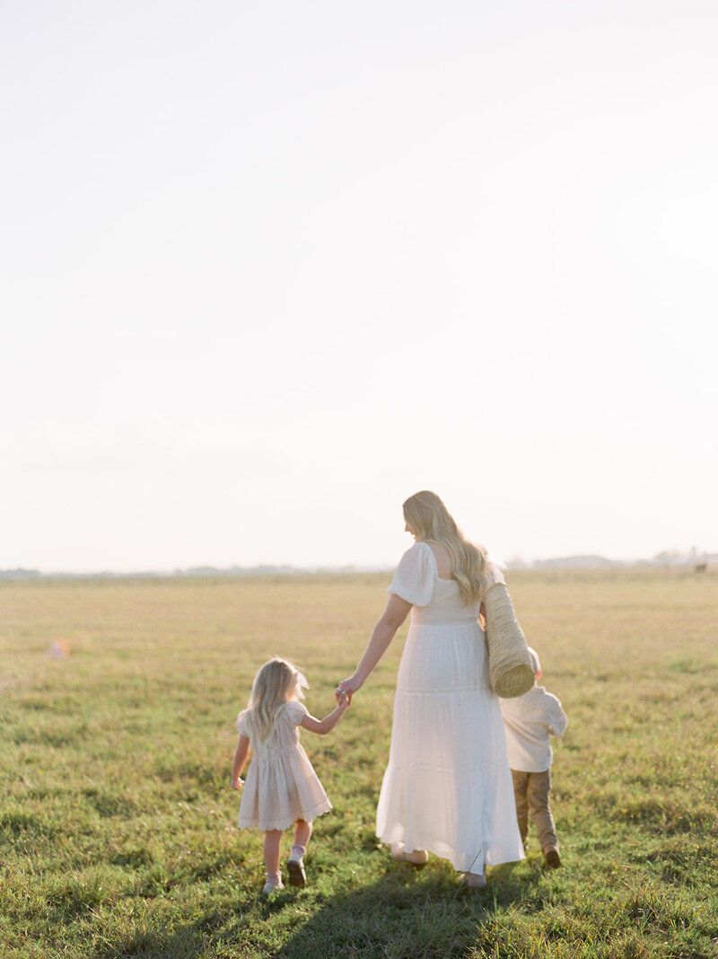 Mother in white dress holds hands with children while walking in sunny meadow.