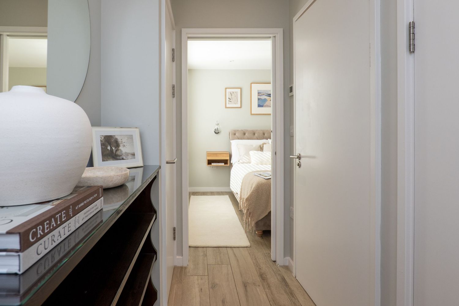 View down a hallway with wood floors and white walls, looking into a bright bedroom through an open doorway.
