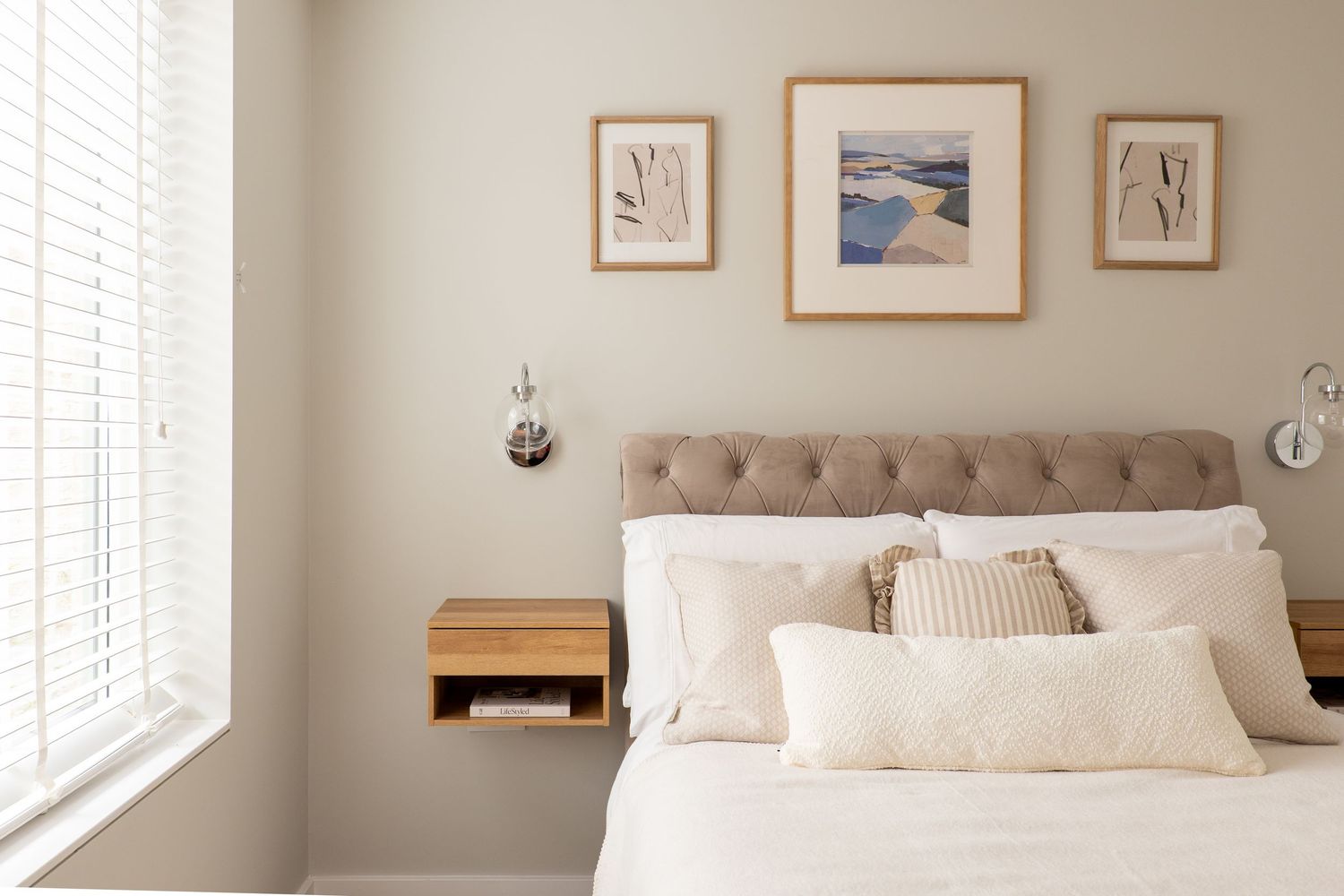 Modern bedroom interior with tufted headboard, framed artwork, and natural light streaming through windows.