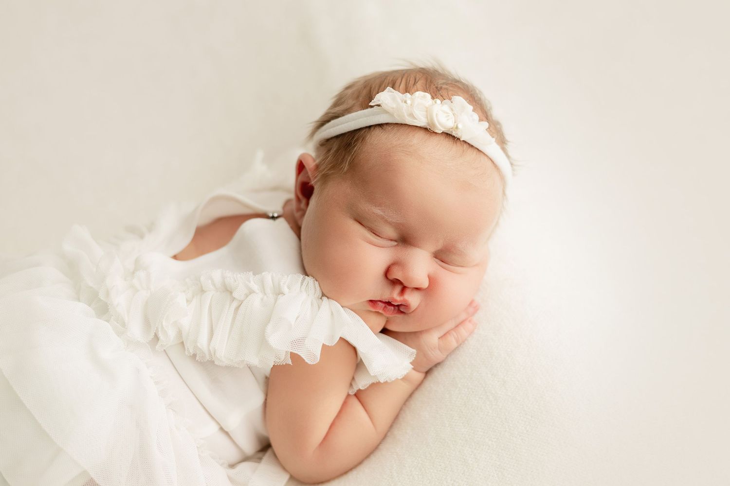 Sleeping newborn baby wearing white lace headband and dress on white backdrop.
