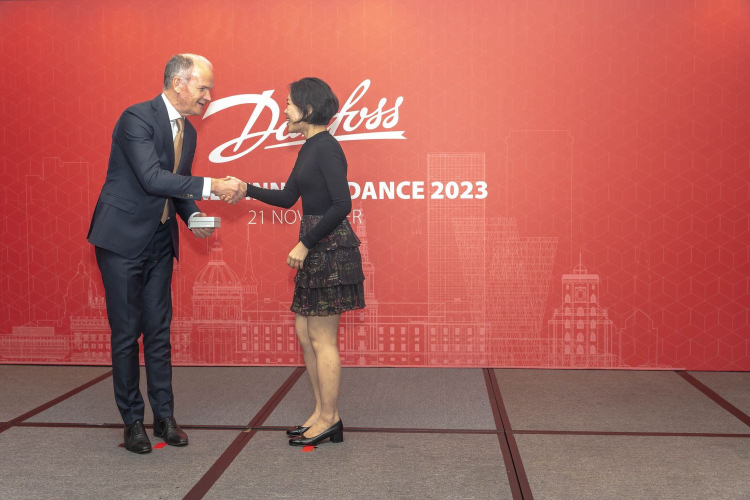 People interacting at formal event with red backdrop featuring cityscape design.