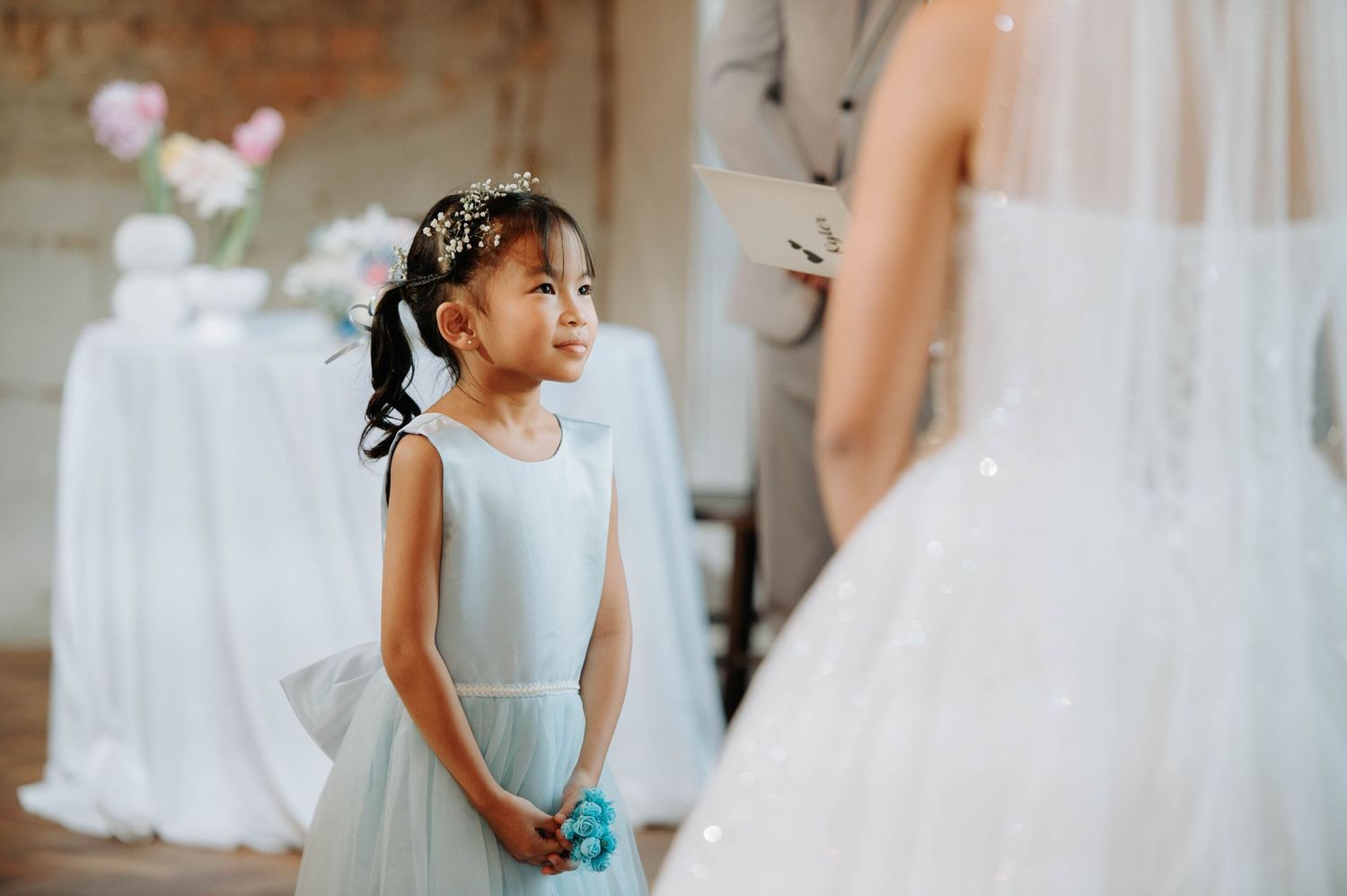 A young attendant in a light blue dress carries a small bouquet during a wedding ceremony.