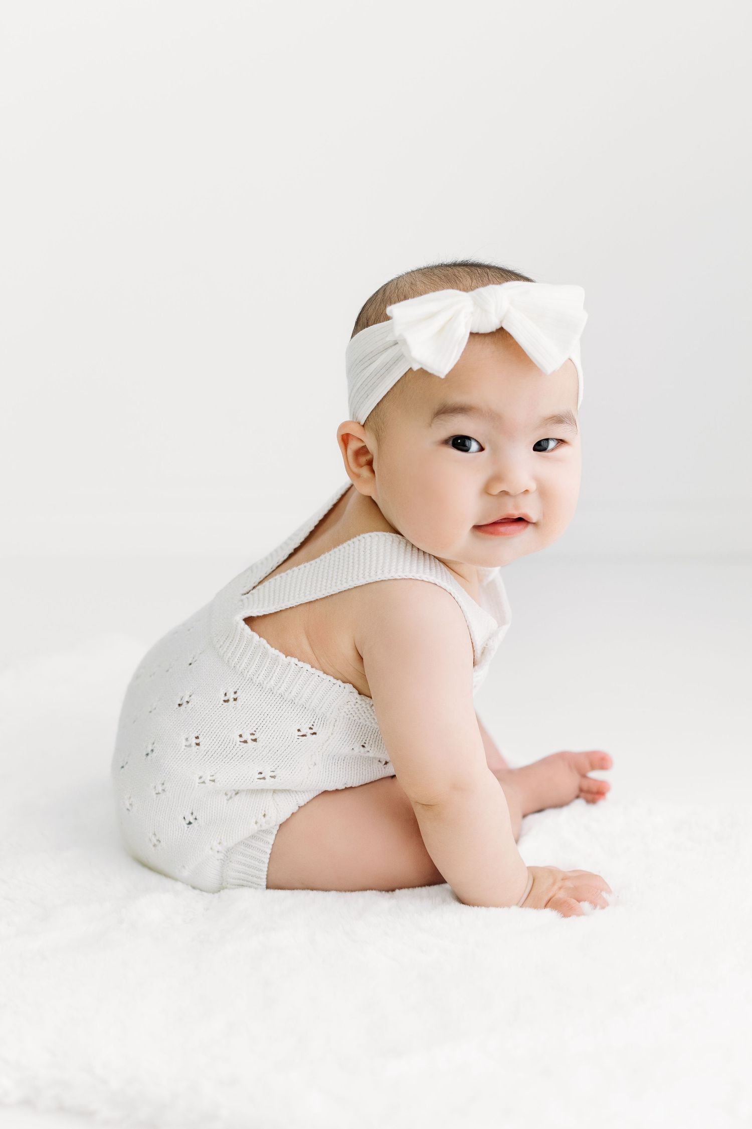 Adorable Asian baby girl, sitting on white rug, smiling, and wearing a white outfit with a big bow.