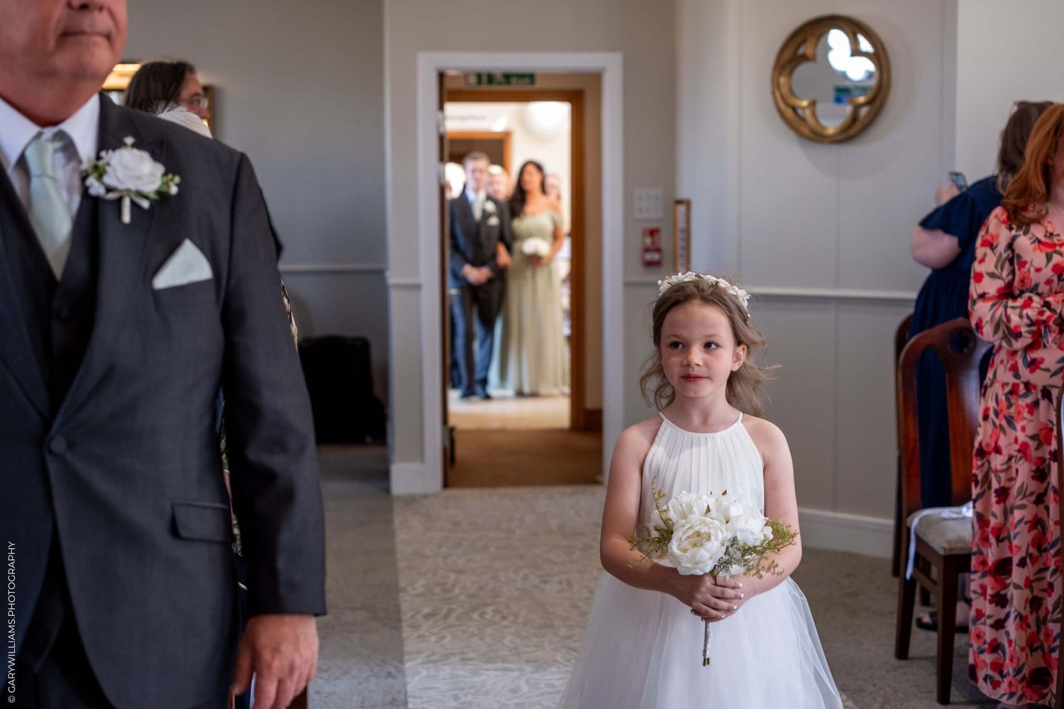 A young attendant in a white dress holds a small bouquet during a wedding ceremony in an elegant interior hallway.