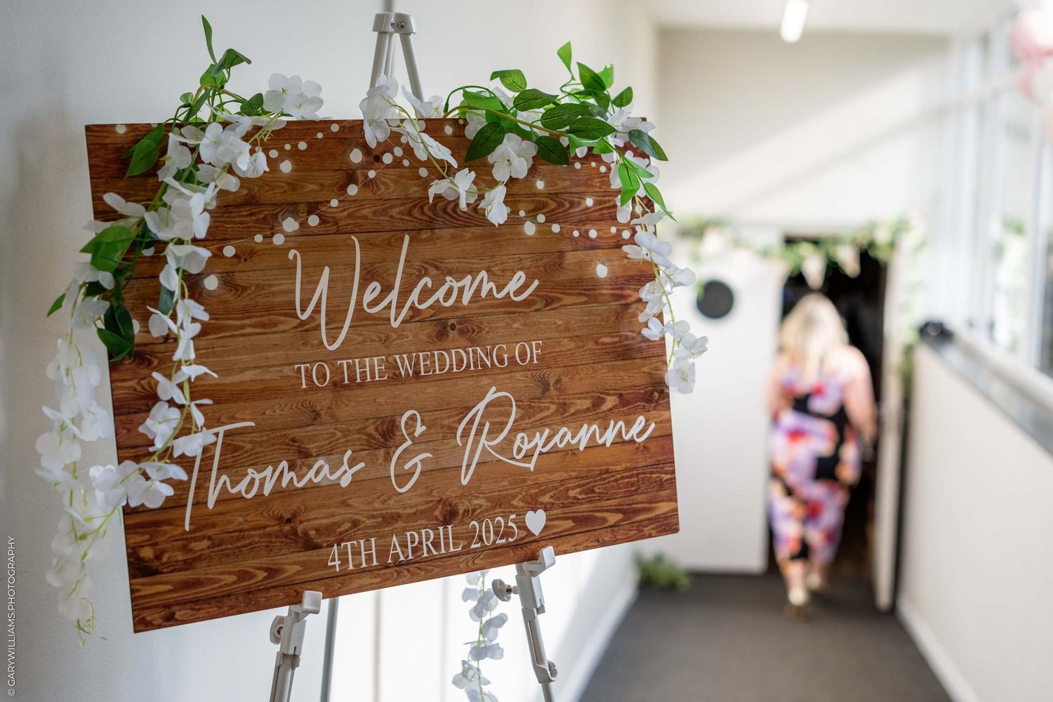 Rustic wooden welcome sign with floral garlands displayed on an easel at a wedding venue.