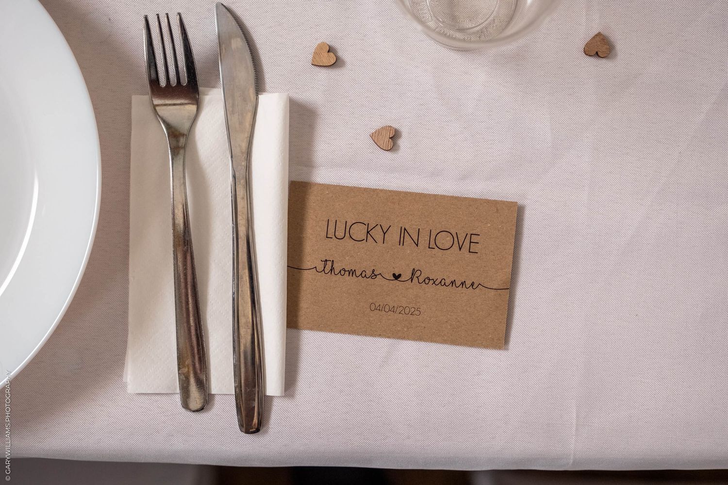 Rustic place setting with silverware and kraft paper place card reading "Lucky in Love" on white tablecloth.