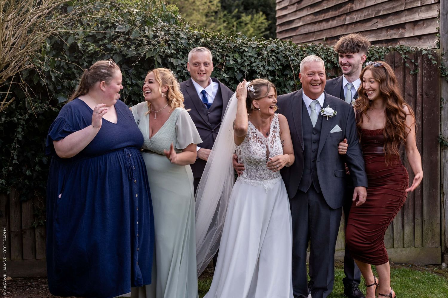Wedding party gathered outside smiling together wearing formal attire in blue, burgundy and mint green dresses.