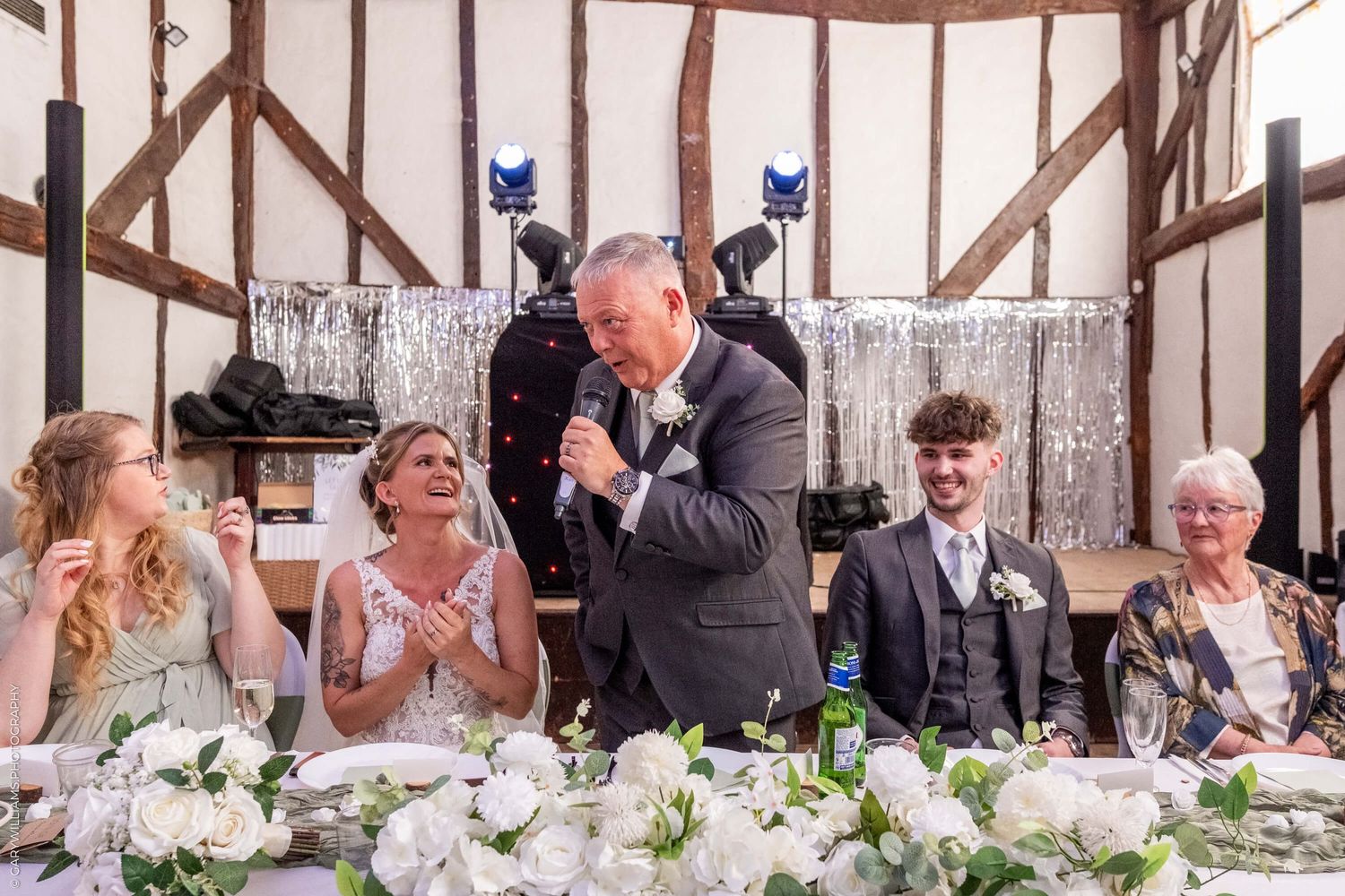 Wedding reception scene with people laughing at a head table decorated with white flowers and silver accents.
