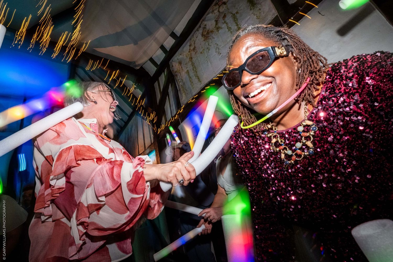 Two people dancing energetically at a party with colorful lighting and glowsticks in a dark venue.