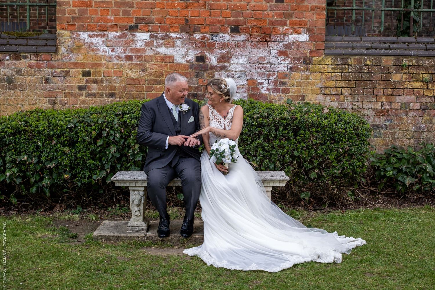 A couple sits together on a stone bench against a rustic brick wall with greenery at a wedding venue.