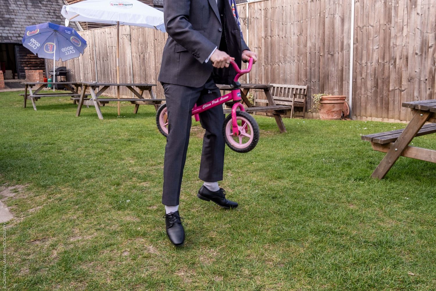 Person in dark suit holds small pink children's bicycle on green lawn next to wooden fence and outdoor furniture.