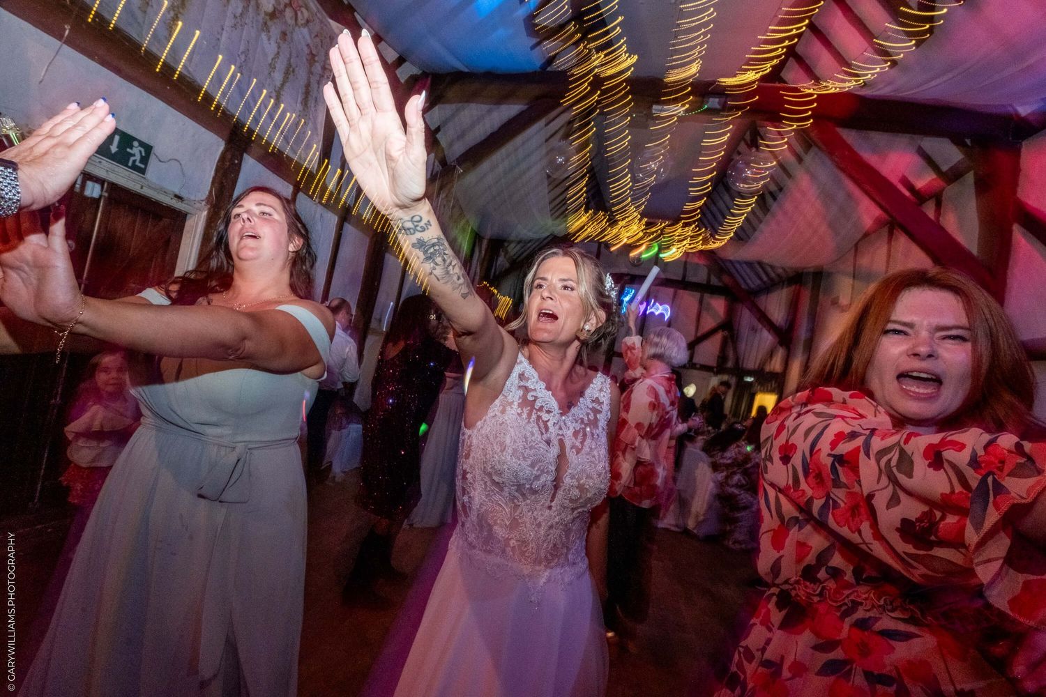 Wedding guests dancing enthusiastically on a dance floor under colorful lighting at an indoor reception venue.