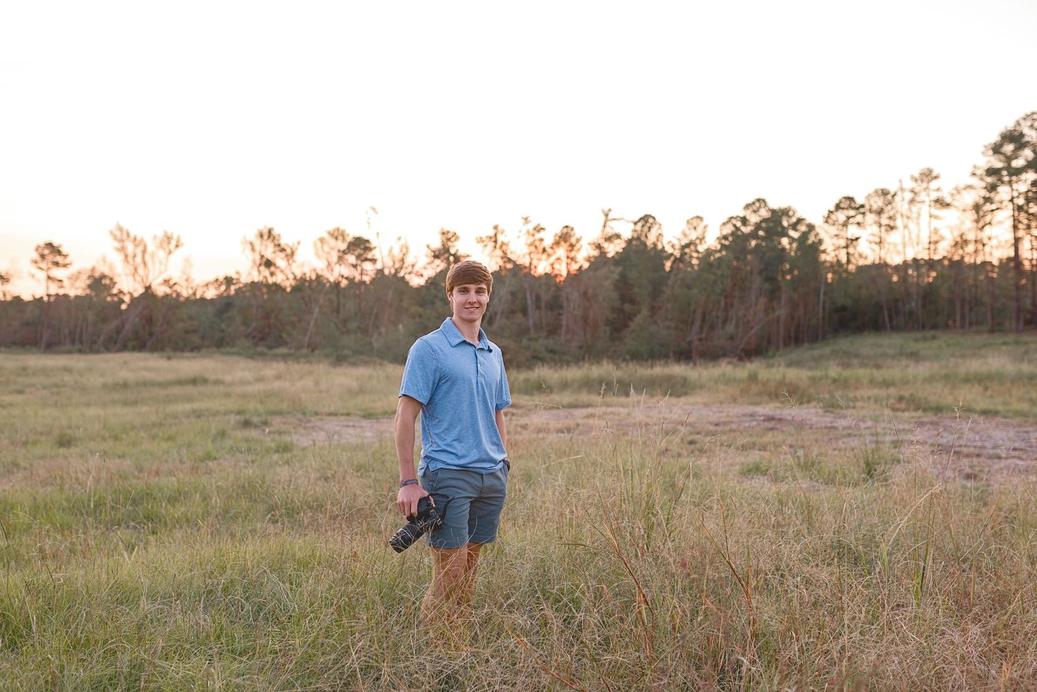 Person carrying photography equipment walks through sunlit meadow at dusk.
