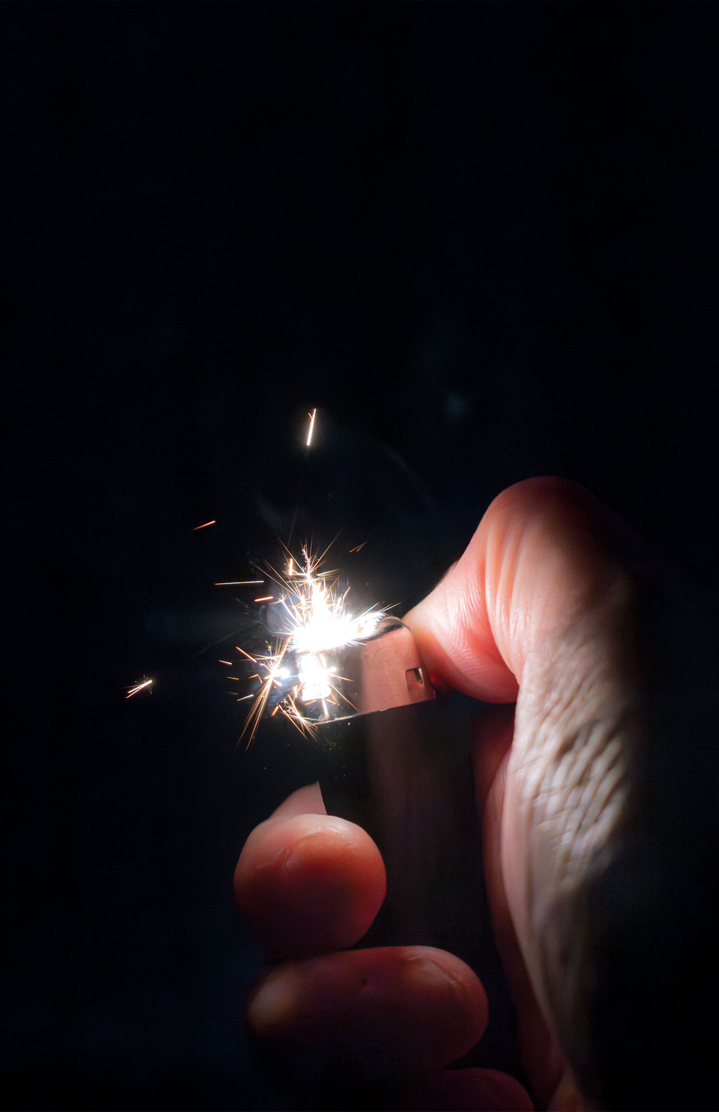 Close-up of a hand holding a sparking lighter against a dark background.