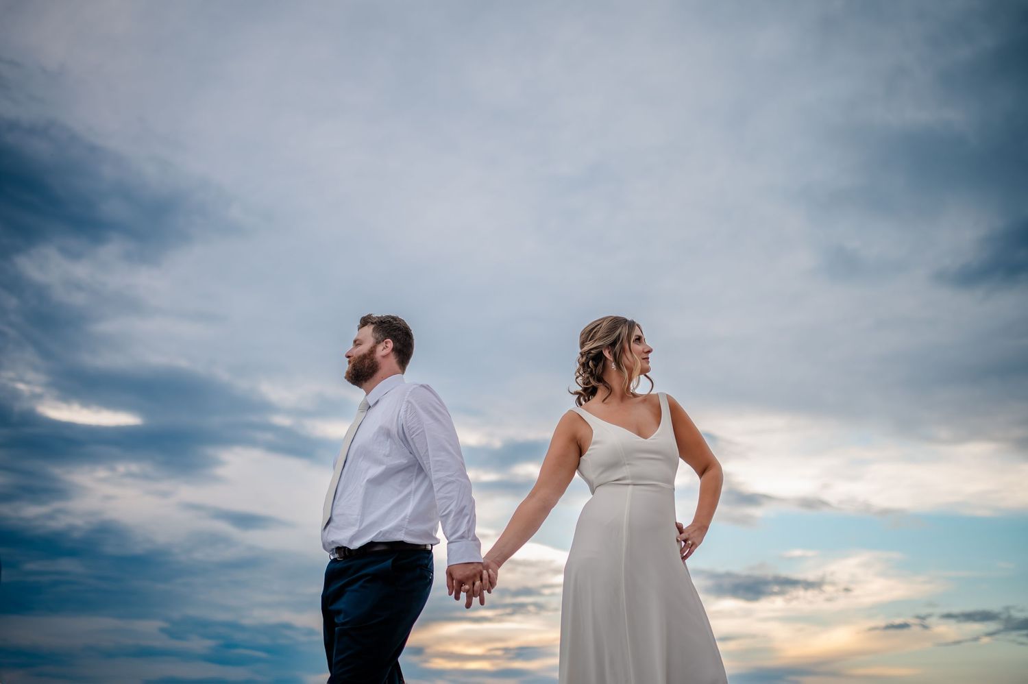 Two people in wedding attire hold hands while looking towards the sunset against a dramatic cloudy sky.