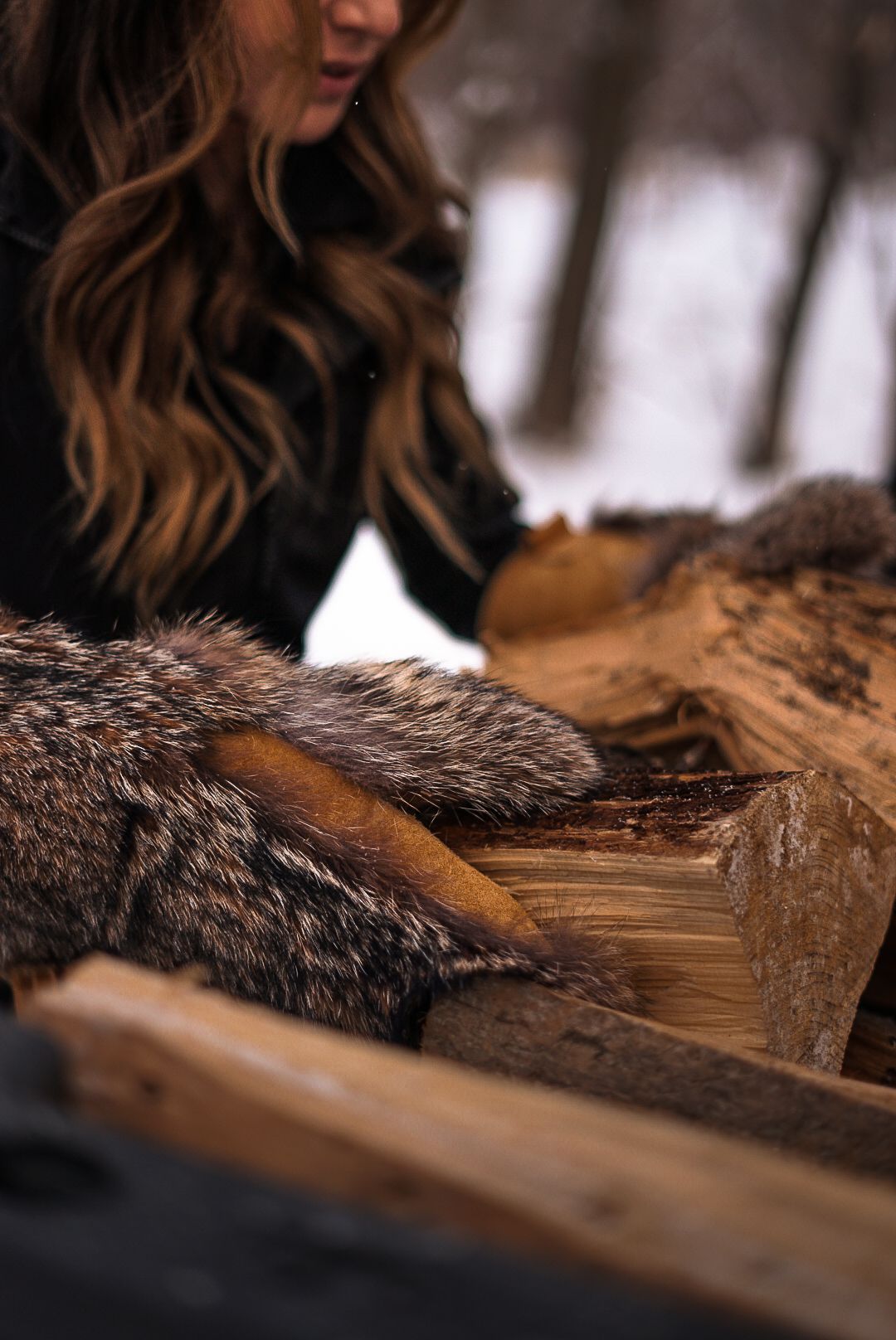 Fur gloves resting on stack of chopped firewood in winter setting.