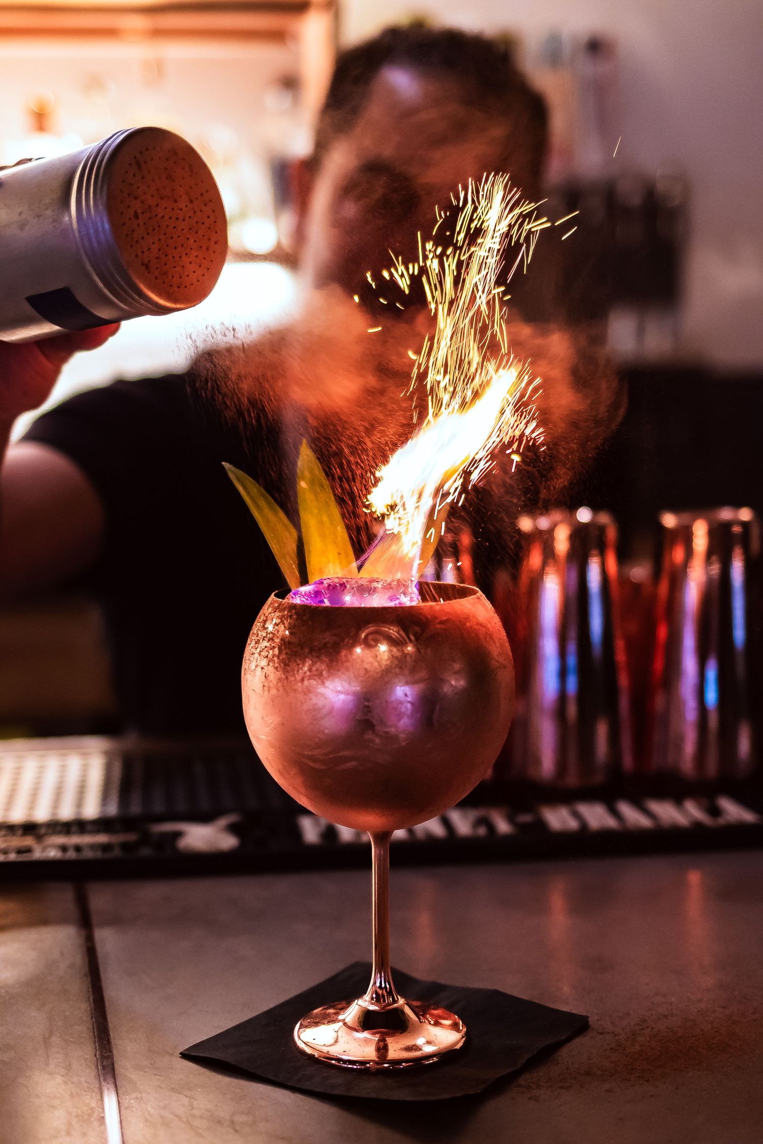 A bartender sprinkles spices onto a flaming cocktail creating dramatic sparks in a dark bar setting.