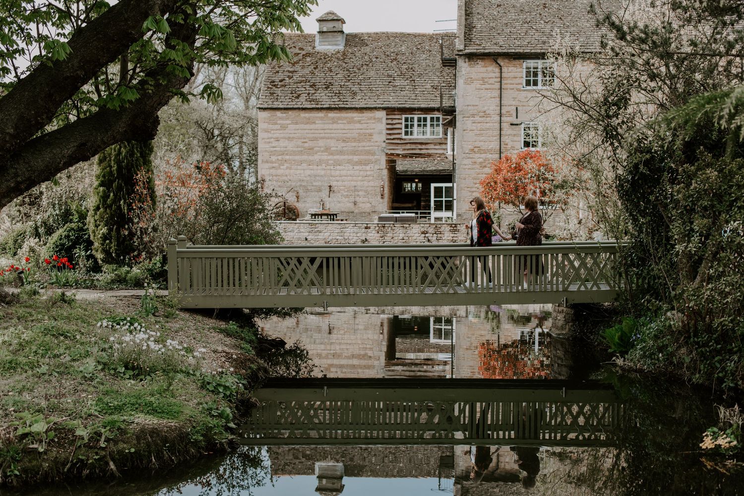 Two ladies are walking across a bridge hand in hand with a water mill wedding venue in the background.