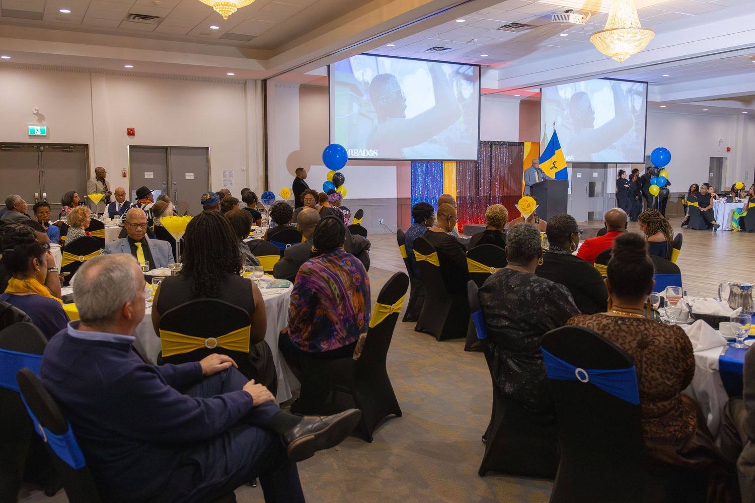 Conference attendees in a large meeting room watch a presentation on dual projection screens with blue balloons decorating the space.