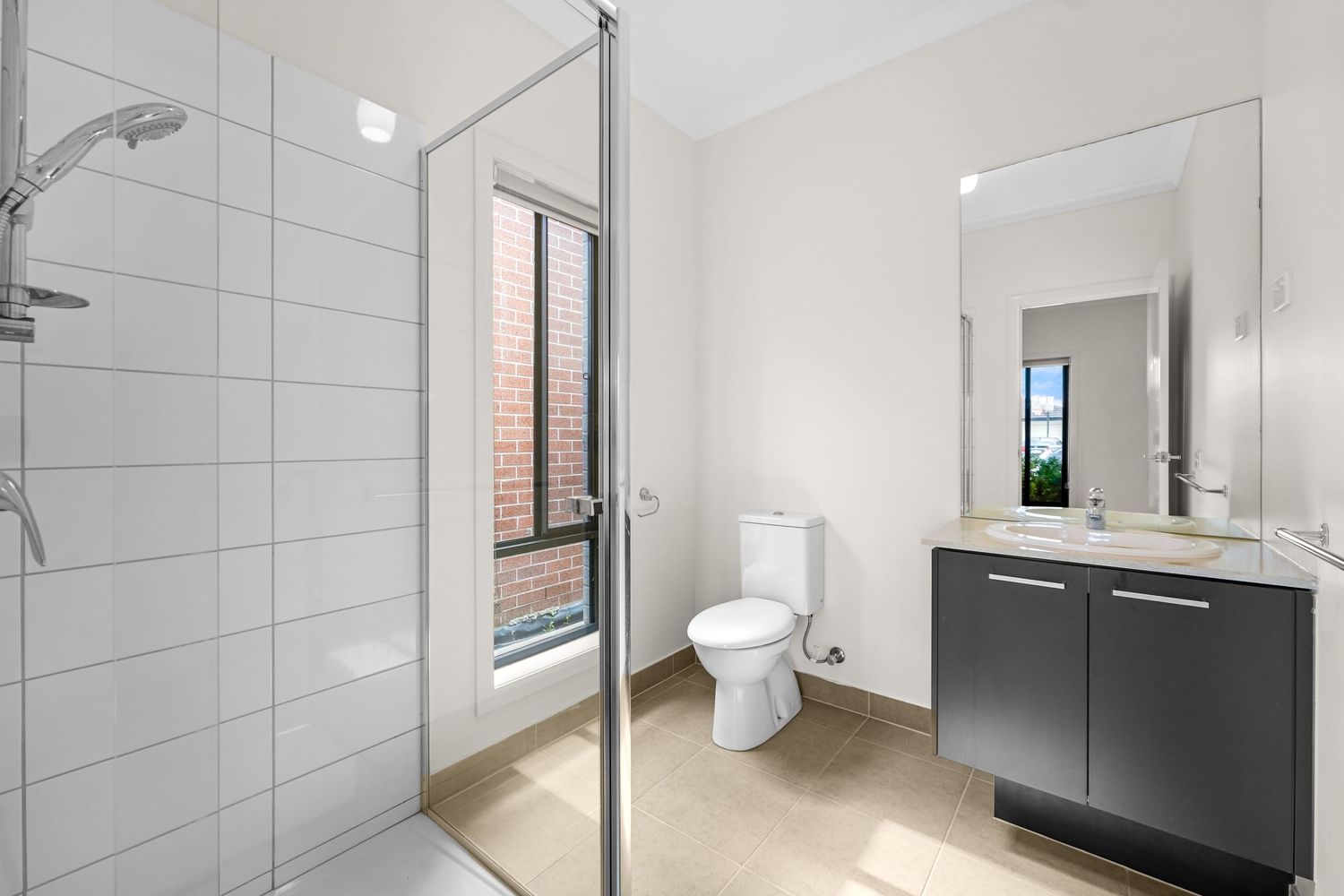 Modern bathroom with gray vanity, white toilet, glass shower enclosure and natural light from window.