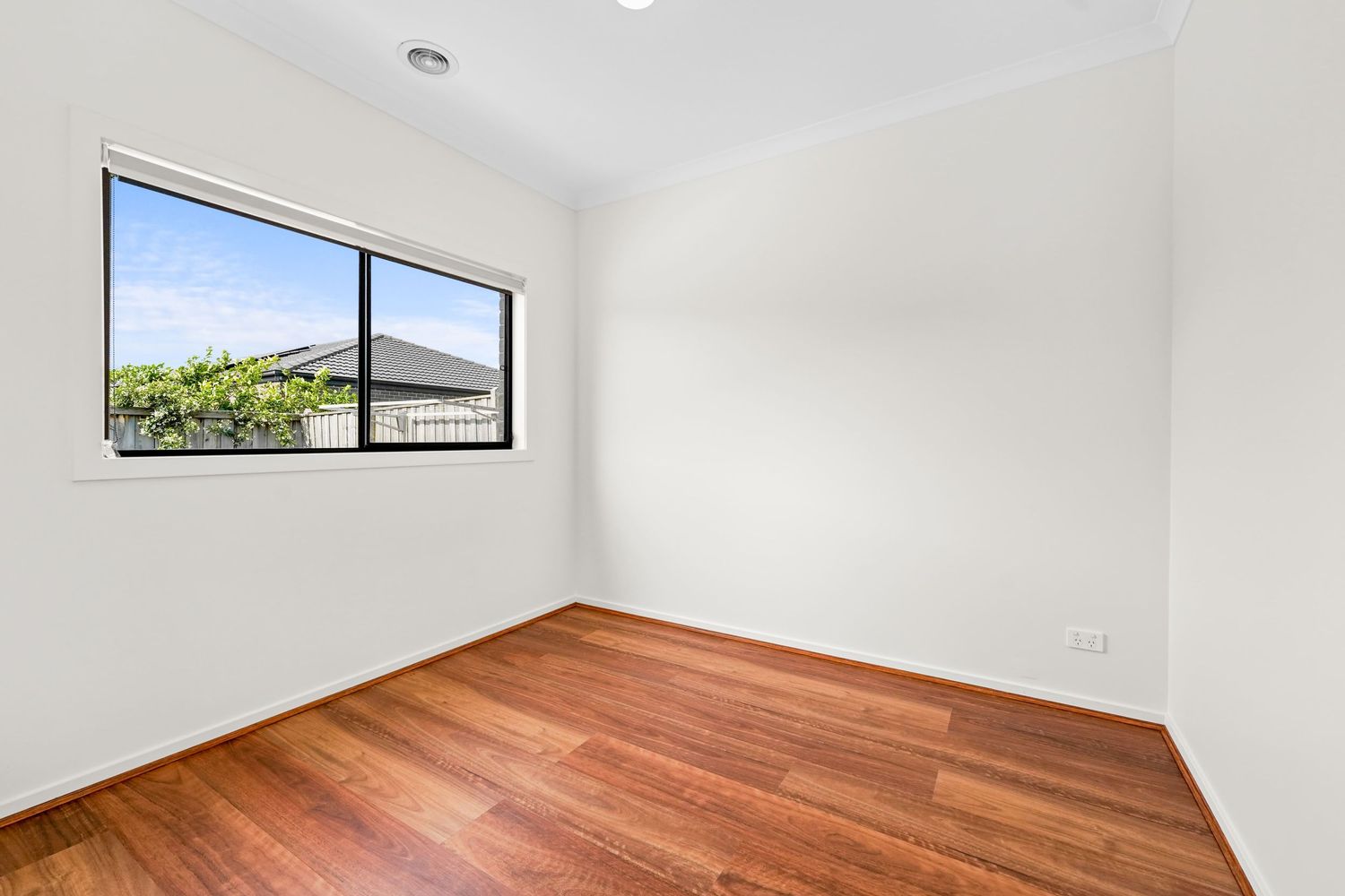 Empty bedroom with polished hardwood floors, white walls, and a window offering views of greenery and neighboring homes.